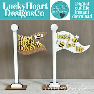 May include: Two wooden flag-shaped signs on stands. The left sign is brown with yellow text reading FARM FRESH HONEY SWEET BUZZING GOODNESS and two bees. The right sign is white with yellow text reading busy bee life and two bees.