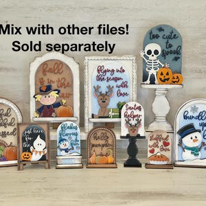 May include: A collection of decorative wooden signs with fall and winter themes. Designs include a scarecrow, reindeer, snowman, ghost, and skeleton, with phrases like "Fall is in the air" and "Too cute to spook".