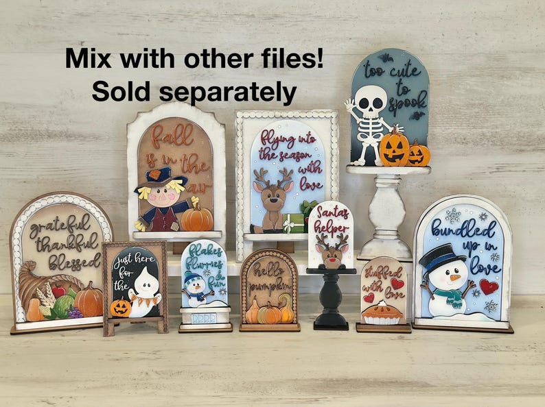 May include: A collection of decorative wooden signs with fall and winter themes. Designs include a scarecrow, reindeer, snowman, ghost, and skeleton, with phrases like "Fall is in the air" and "Too cute to spook".