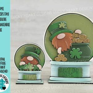 Könnte beinhalten: Zwei Schneekugel-Ornamente aus Holz mit einem Gnom, der einen grünen Hut trägt und einen Topf mit Gold hält. Die größere Schneekugel hat den Text "Lucky and I Gnome It" auf dem Sockel. Die kleinere Schneekugel hat den Text "Lucky" auf dem Sockel.