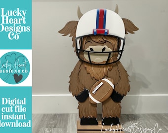 May include: A layered wooden cutout of a brown highland cow wearing a white football helmet with a red and blue center stripe and a black face mask. The cow is holding a brown football with white laces. Text reads Lucky Heart Designs Co.
