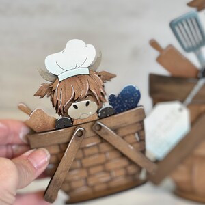 May include: A wooden craft of a Highland cow wearing a chef's hat, sitting in a brown wicker basket with a blue and white patterned fabric inside. The basket has two wooden handles.
