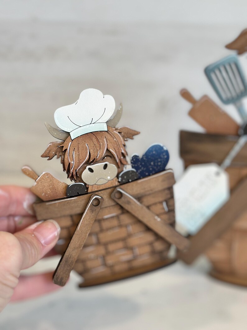 May include: A wooden craft of a Highland cow wearing a chef's hat, sitting in a brown wicker basket with a blue and white patterned fabric inside. The basket has two wooden handles.