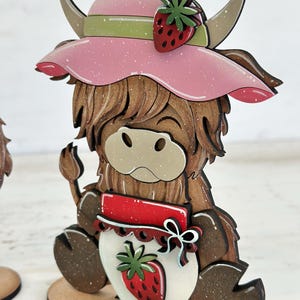 May include: A wooden figurine of a Highland cow wearing a pink hat with a strawberry on top. The cow is holding a jar with a strawberry on the front. The figurine is painted in brown, white, red, and green.