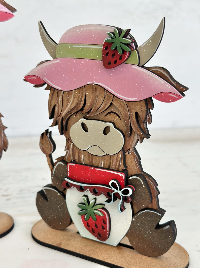 May include: A wooden figurine of a Highland cow wearing a pink hat with a strawberry on top. The cow is holding a jar with a strawberry on the front. The figurine is painted in brown, white, red, and green.