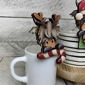 May include: A wooden reindeer mug hugger with a candy cane. The reindeer has brown antlers, a light brown face, and a fluffy, light brown mane. The candy cane is red and white. The mug is white.