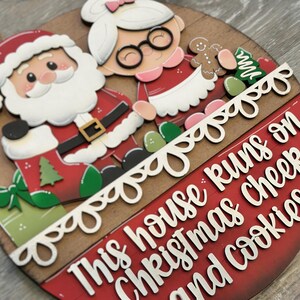 Puede incluir: Decoraci&oacute;n navide&ntilde;a redonda de madera con Pap&aacute; Noel y Mam&aacute; Noel. El dise&ntilde;o incluye una pancarta roja y blanca con el texto "This house runs on Christmas cheer and cookies."