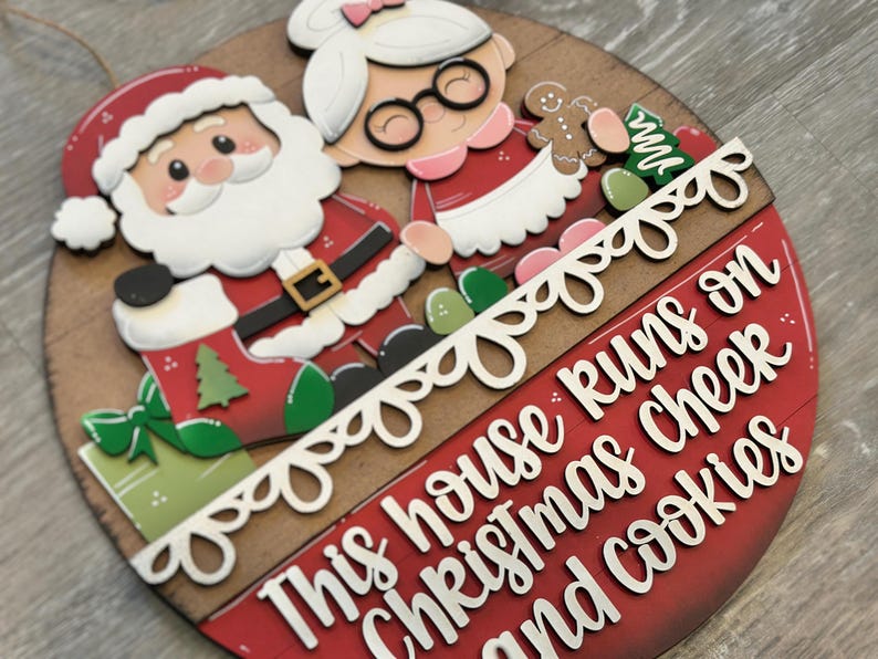 Puede incluir: Decoraci&oacute;n navide&ntilde;a redonda de madera con Pap&aacute; Noel y Mam&aacute; Noel. El dise&ntilde;o incluye una pancarta roja y blanca con el texto "This house runs on Christmas cheer and cookies."
