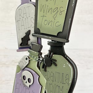 May include: Wooden Halloween potion bottles with labels reading "Rat Tails," "Bat Wings Tonic," "Arsenic Poison," and "Tarantula Venom." The bottles are in shades of purple, green, and black, with a rat, skull, and spider embellishments.