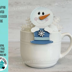 May include: White ceramic mug with a snowman mug topper. The snowman is white with a blue hat and a carrot nose. The mug is sitting on a white surface.