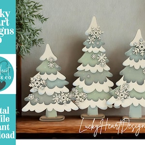 May include: Three wooden Christmas tree decorations with a layered design, painted in white, light blue, and green. Each tree is adorned with snowflake accents. The image includes the text "Lucky Heart Designs Co" and "Digital cut file instant download".