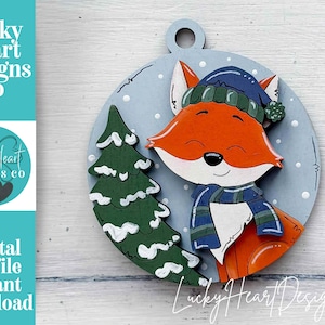 May include: A wooden Christmas ornament featuring a cartoon fox wearing a blue hat and scarf, with a green Christmas tree on a light blue background. The ornament has a loop for hanging and the text "Lucky Heart Designs Co" is visible.