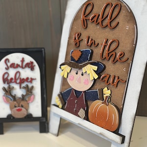 May include: Wooden decorative signs with a fall theme. One sign features a scarecrow and pumpkin with the text "Fall is in the air." The other sign has a reindeer and the text "Santa's Helper."