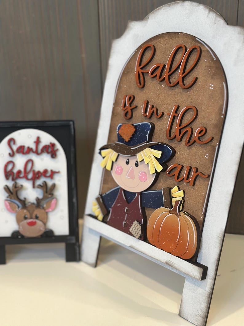 May include: Wooden decorative signs with a fall theme. One sign features a scarecrow and pumpkin with the text "Fall is in the air." The other sign has a reindeer and the text "Santa's Helper."