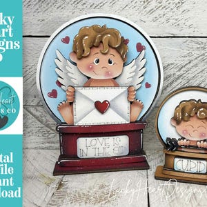 May include: Wooden Valentine's Day decorations featuring a cherub holding an envelope with a heart. The larger piece has the text "LOVE IS IN THE AIR". The smaller piece says "CUPID". The background is light blue with red hearts.