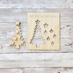 Puede incluir: Un kit de adornos de &aacute;rbol de Navidad de madera con un recorte de un &aacute;rbol y adornos. El texto "Build your own tree ornament" est&aacute; escrito en la madera.