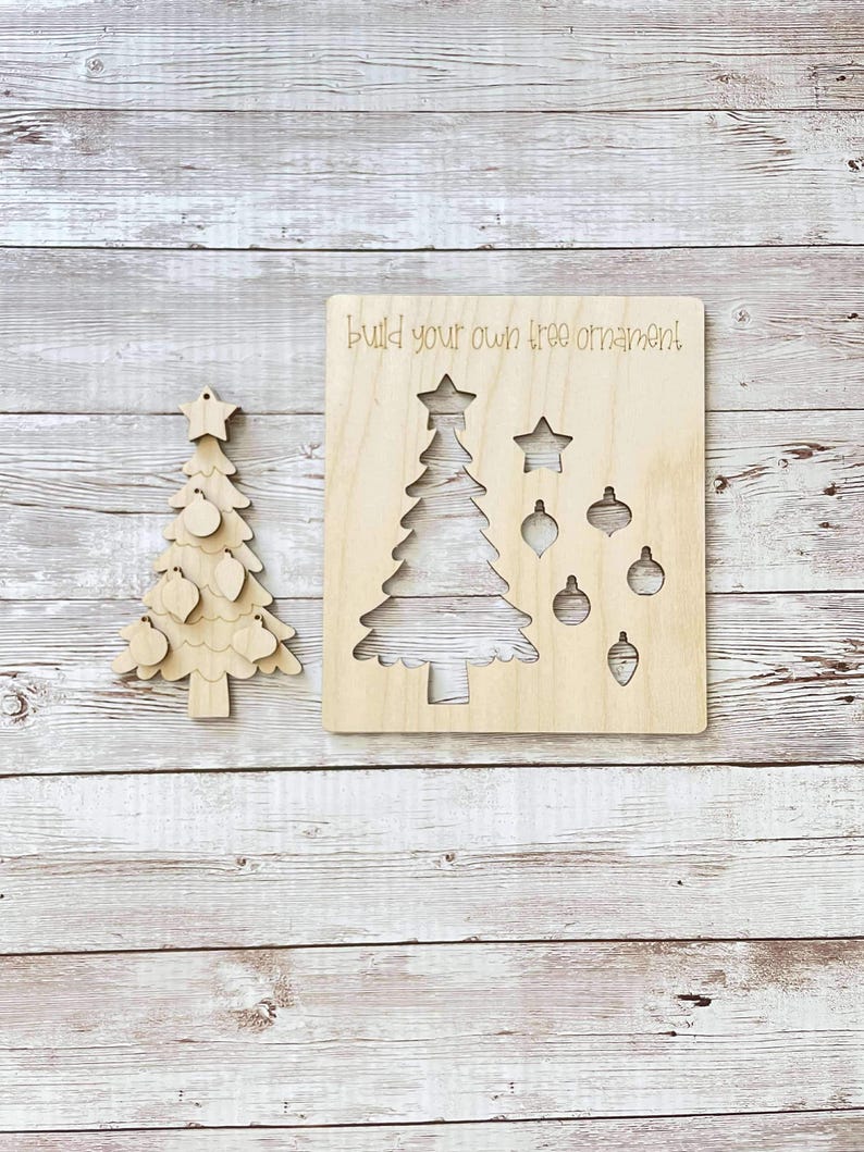 Puede incluir: Un kit de adornos de &aacute;rbol de Navidad de madera con un recorte de un &aacute;rbol y adornos. El texto "Build your own tree ornament" est&aacute; escrito en la madera.