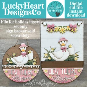 Puede incluir: Dos letreros de madera con temática de gansos. Uno es redondo, el otro rectangular. Ambos presentan un ganso blanco con sombrero y lazo rosa, rodeado de flores y el texto "Hey There Silly Goose". Los letreros son para un juego de inserción navideña.