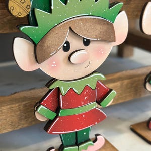 May include: Wooden Christmas elf decoration. The elf has a green hat and collar, a red tunic, and green pants. The elf's face is light-skinned with rosy cheeks and a small smile. The elf is standing on a small wooden base.