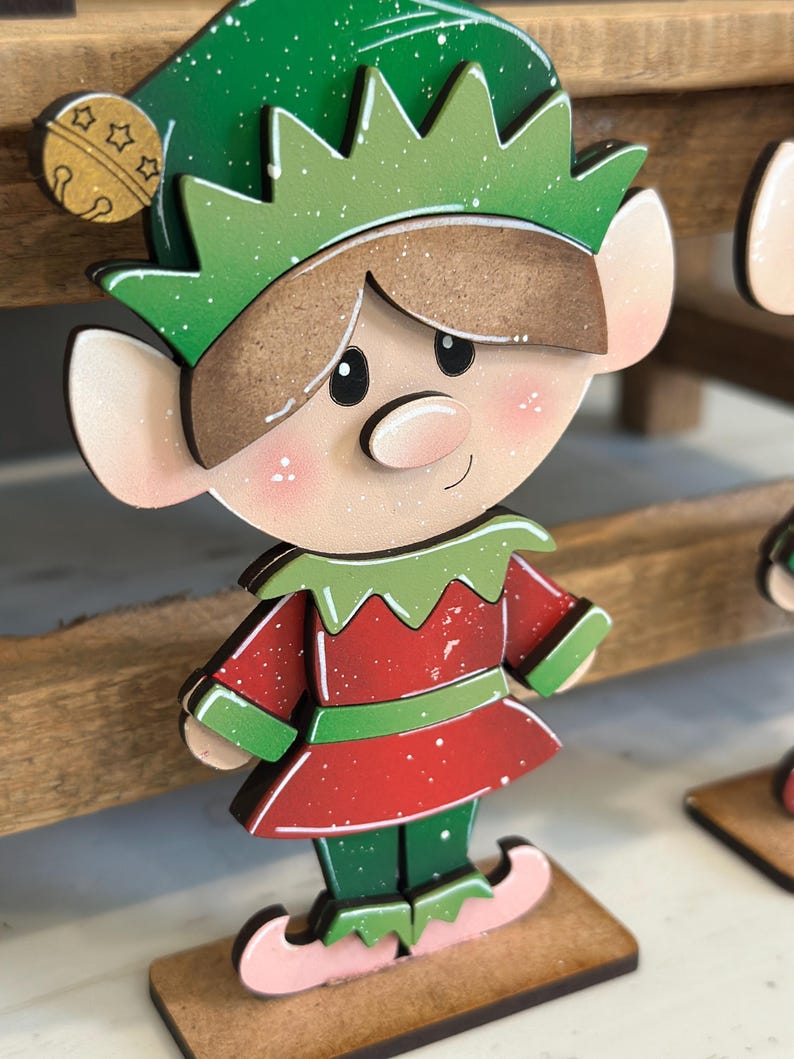 May include: Wooden Christmas elf decoration. The elf has a green hat and collar, a red tunic, and green pants. The elf's face is light-skinned with rosy cheeks and a small smile. The elf is standing on a small wooden base.
