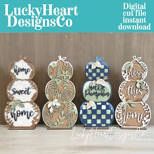May include: Four stacked pumpkin-shaped wooden signs with laser-cut designs. The first features 'home sweet home' text, the second has floral patterns, the third displays 'hello pumpkin' with a blue plaid, and the fourth reads 'Bless this home'.