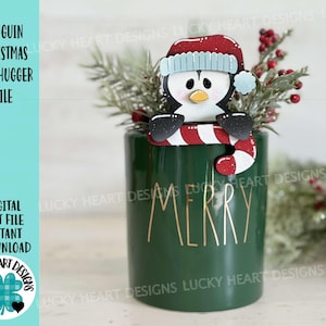May include: A green ceramic mug with the word "Merry" written in gold lettering. A wooden penguin wearing a red Santa hat and holding a candy cane is perched on the rim of the mug.