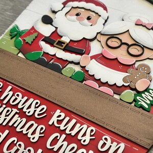 Puede incluir: Decoraci&oacute;n navide&ntilde;a de madera con Pap&aacute; Noel y Mam&aacute; Noel. La decoraci&oacute;n tiene el texto "This house runs on Christmas and cocoa". La combinaci&oacute;n de colores incluye rojo, blanco, verde y marr&oacute;n. Las figuras son de estilo festivo.