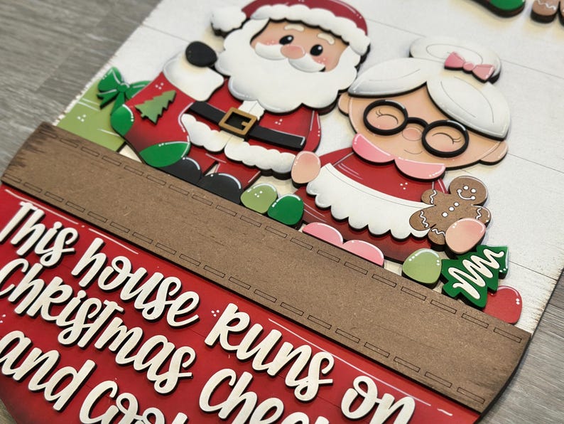 Puede incluir: Decoraci&oacute;n navide&ntilde;a de madera con Pap&aacute; Noel y Mam&aacute; Noel. La decoraci&oacute;n tiene el texto "This house runs on Christmas and cocoa". La combinaci&oacute;n de colores incluye rojo, blanco, verde y marr&oacute;n. Las figuras son de estilo festivo.