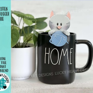May include: A gray cat kitten mug hugger holding a blue ball of yarn. The mug hugger is sitting on a black mug with the word "HOME" written in white on the front.