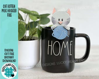 May include: A gray cat kitten mug hugger holding a blue ball of yarn. The mug hugger is sitting on a black mug with the word "HOME" written in white on the front.