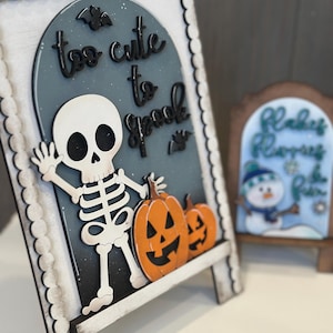 May include: A wooden Halloween-themed sign with a skeleton, pumpkins, and the phrase "too cute to spook." The sign is white, black, and orange, with a scalloped border. A smaller sign with a snowman is in the background.