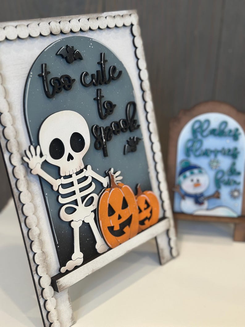 May include: A wooden Halloween-themed sign with a skeleton, pumpkins, and the phrase "too cute to spook." The sign is white, black, and orange, with a scalloped border. A smaller sign with a snowman is in the background.