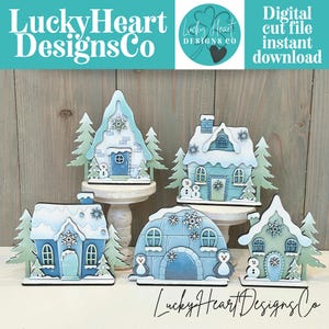 May include: A collection of five winter-themed wooden house cutouts. Each house is painted in shades of blue and white, with snow-covered roofs and decorative trees. The text "Lucky Heart Designs Co" is visible.