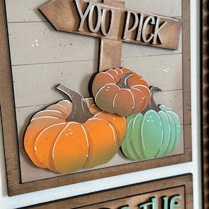 Peut inclure: Panneau en bois avec les mots "YOU PICK" au-dessus de trois citrouilles dans des tons orange et vert. Les citrouilles sont plac&eacute;es sur un fond brun clair avec un cadre en bois. Le panneau est une d&eacute;coration sur le th&egrave;me de l'automne.
