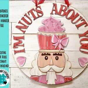 May include: A round wooden door hanger with a pink and red nutcracker design. The nutcracker is wearing a pink cupcake hat with a pink bow. The text "I'm nuts about you" is written around the edge of the hanger.