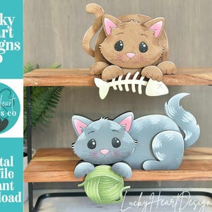 May include: Two wooden cat cutouts on a wooden shelf. The top cat is brown and holding a fish bone. The bottom cat is gray and holding a green ball of yarn. The text "Lucky Heart Designs Co" and "Digital cut file instant download" are visible.