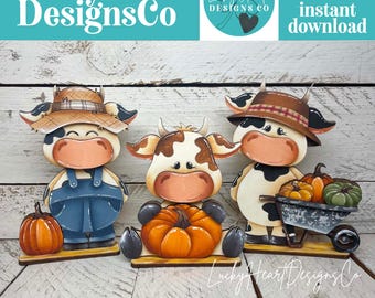 May include: Three wooden cow cutouts with fall themes. The left cow wears a straw hat and blue overalls with a pumpkin. The center cow holds a pumpkin. The right cow wears a brown hat and pushes a wheelbarrow filled with orange and green pumpkins.