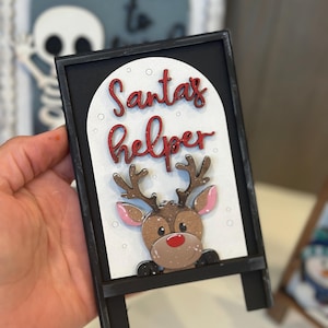 May include: A black framed sign with the words "Santa's helper" in red script. The sign features a cartoon reindeer with brown antlers, a red nose, and pink ears. The sign is a Christmas decoration.