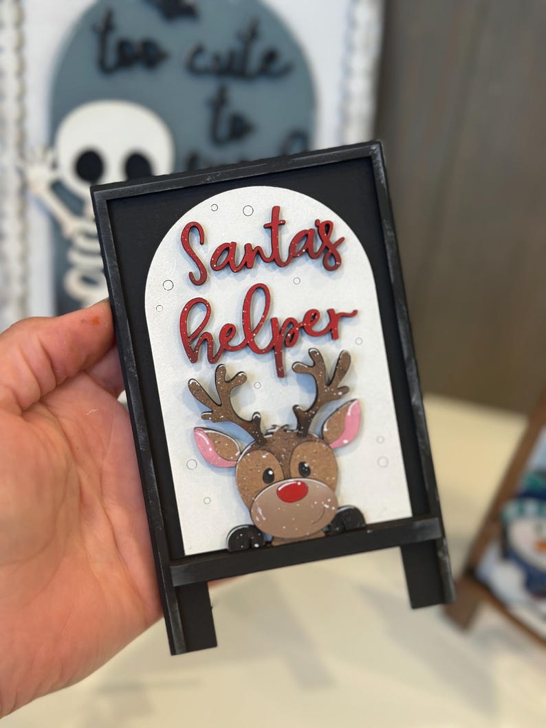 May include: A black framed sign with the words "Santa's helper" in red script. The sign features a cartoon reindeer with brown antlers, a red nose, and pink ears. The sign is a Christmas decoration.