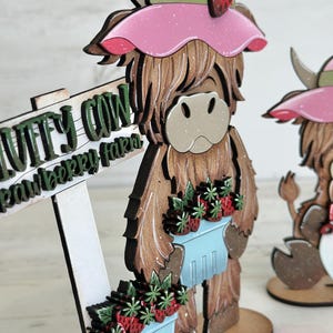 May include: A wooden figurine of a Highland cow wearing a pink hat with a green ribbon and a strawberry on top. The cow is holding a blue basket filled with red strawberries. The cow is standing on a wooden base with a sign that reads "Wuffy Cow Strawberry Farm".