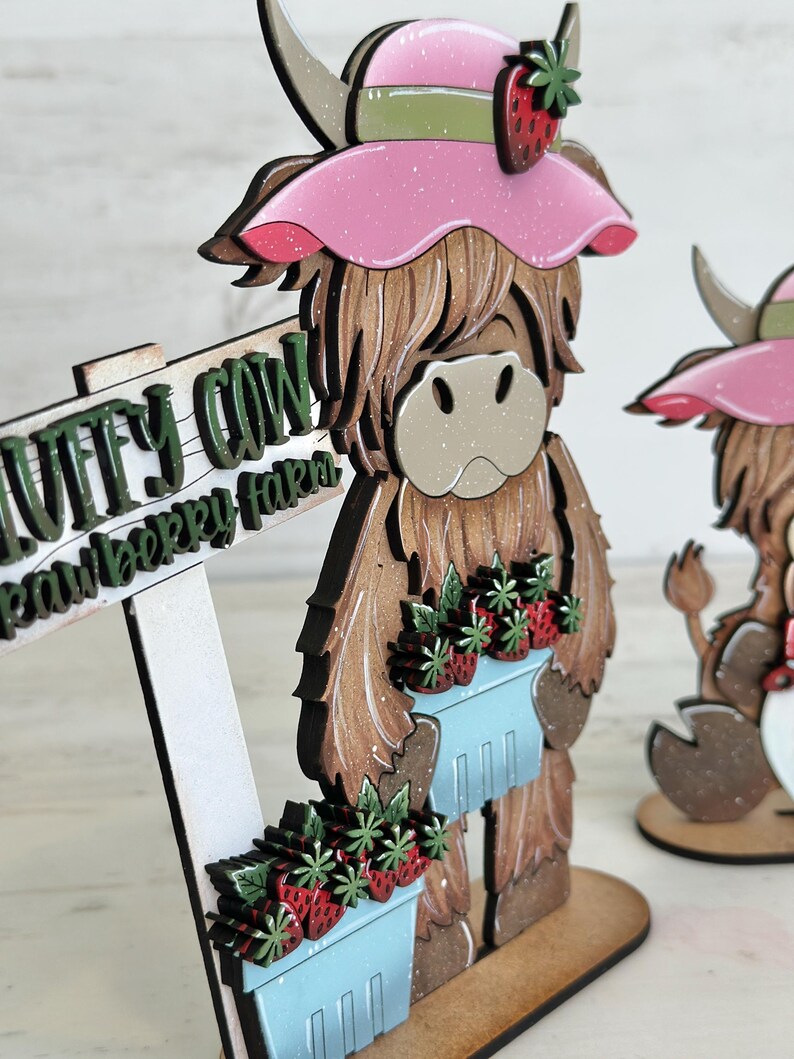 May include: A wooden figurine of a Highland cow wearing a pink hat with a green ribbon and a strawberry on top. The cow is holding a blue basket filled with red strawberries. The cow is standing on a wooden base with a sign that reads "Wuffy Cow Strawberry Farm".