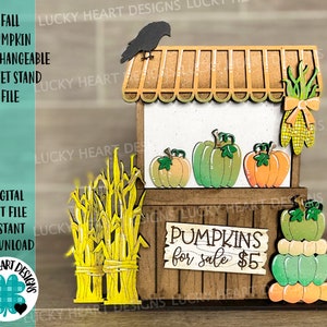 以下が含まれることがあります： 「かぼちゃ販売中 $5」と書かれた木製の秋の南瓜のマーケットスタンド。スタンドはトウモロコシの穂軸、南瓜、黒いカラスで飾られています。