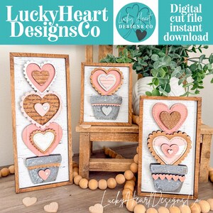 May include: Three framed wooden heart decorations. Each features a pink heart design with a scalloped edge, layered over a gray pot. The text "Lucky Heart Designs Co" is visible. The background is white with a distressed wood texture.