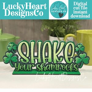 May include: A green and white wooden sign with the phrase "Shake your shamrocks" in a playful font, decorated with shamrock accents. The sign is a digital cut file instant download.