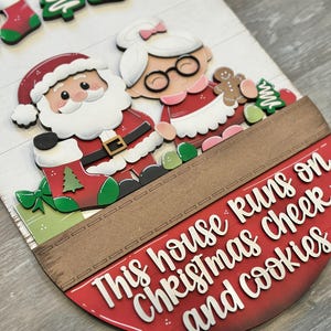 Puede incluir: Un letrero de madera con tem&aacute;tica navide&ntilde;a con Pap&aacute; Noel y Mam&aacute; Noel, hombres de jengibre y adornos de &aacute;rboles de Navidad. El letrero dice: "This house runs on Christmas cheer and cookies."
