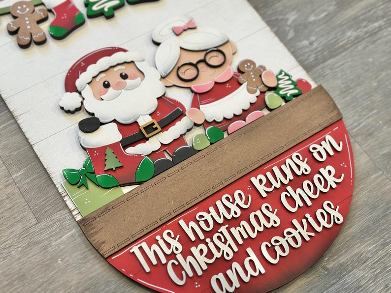 Puede incluir: Un letrero de madera con tem&aacute;tica navide&ntilde;a con Pap&aacute; Noel y Mam&aacute; Noel, hombres de jengibre y adornos de &aacute;rboles de Navidad. El letrero dice: "This house runs on Christmas cheer and cookies."