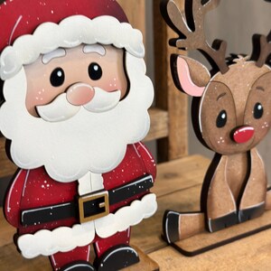 May include: Wooden Christmas decorations featuring Santa Claus and a reindeer. Santa is in a red suit with white trim and a black belt. The reindeer is brown with antlers and a red nose. Both are on wooden bases.