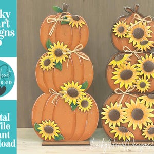 Puede incluir: Dos decoraciones de calabazas apiladas con calabazas naranjas adornadas con girasoles amarillos y lazos de cuerda color canela. La pila izquierda tiene tres calabazas y la derecha dos. El texto Lucky Heart Designs Co es visible.