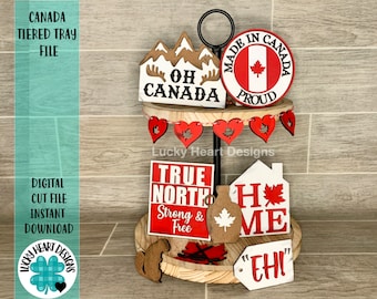 Puede incluir: Una bandeja de niveles decorada con letreros de madera con temática canadiense. El nivel superior presenta un letrero redondo que dice "Made in Canada Proud" con una bandera canadiense en el centro. El nivel intermedio tiene una pancarta con corazones rojos y un letrero que dice "Oh Canada". El nivel inferior tiene un letrero que dice "True North Strong & Free", un letrero que dice "Home" con una hoja de arce, y un letrero que dice "Eh!"