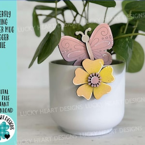 Peut inclure: Une tasse en céramique blanche avec un papillon rose et un motif de fleur jaune. La tasse est posée sur une surface blanche. Le texte "LUCKY HEART DESIGNS" est visible sur la surface.